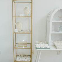 Load image into Gallery viewer, A tall, narrow gold-framed shelving unit with glass shelves stands in a bright room next to a white table and white arched shelf. The shelves display jewelry items, necklaces, and small accessories.
