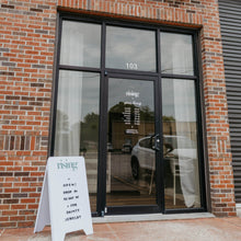 Load image into Gallery viewer, Rising Jewelry storefront on a brick building with a teal sign above black-framed glass doors and a white A-frame sidewalk sign in front.
