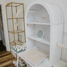 Load image into Gallery viewer, A white arched jewelry display shelf with multiple tiers and a slanted front tray, shown alongside a gold-framed shelving unit in a bright retail or boutique interior with wood flooring.
